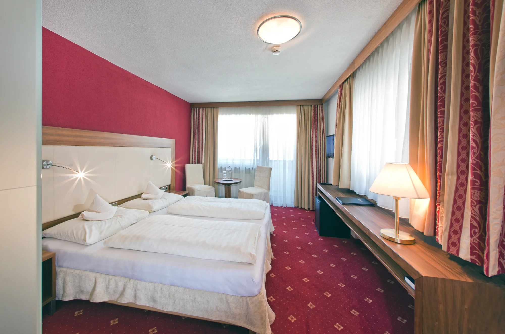Hotelzimmer mit Doppelbett, rotem Teppichboden und Sitzecke am Fenster