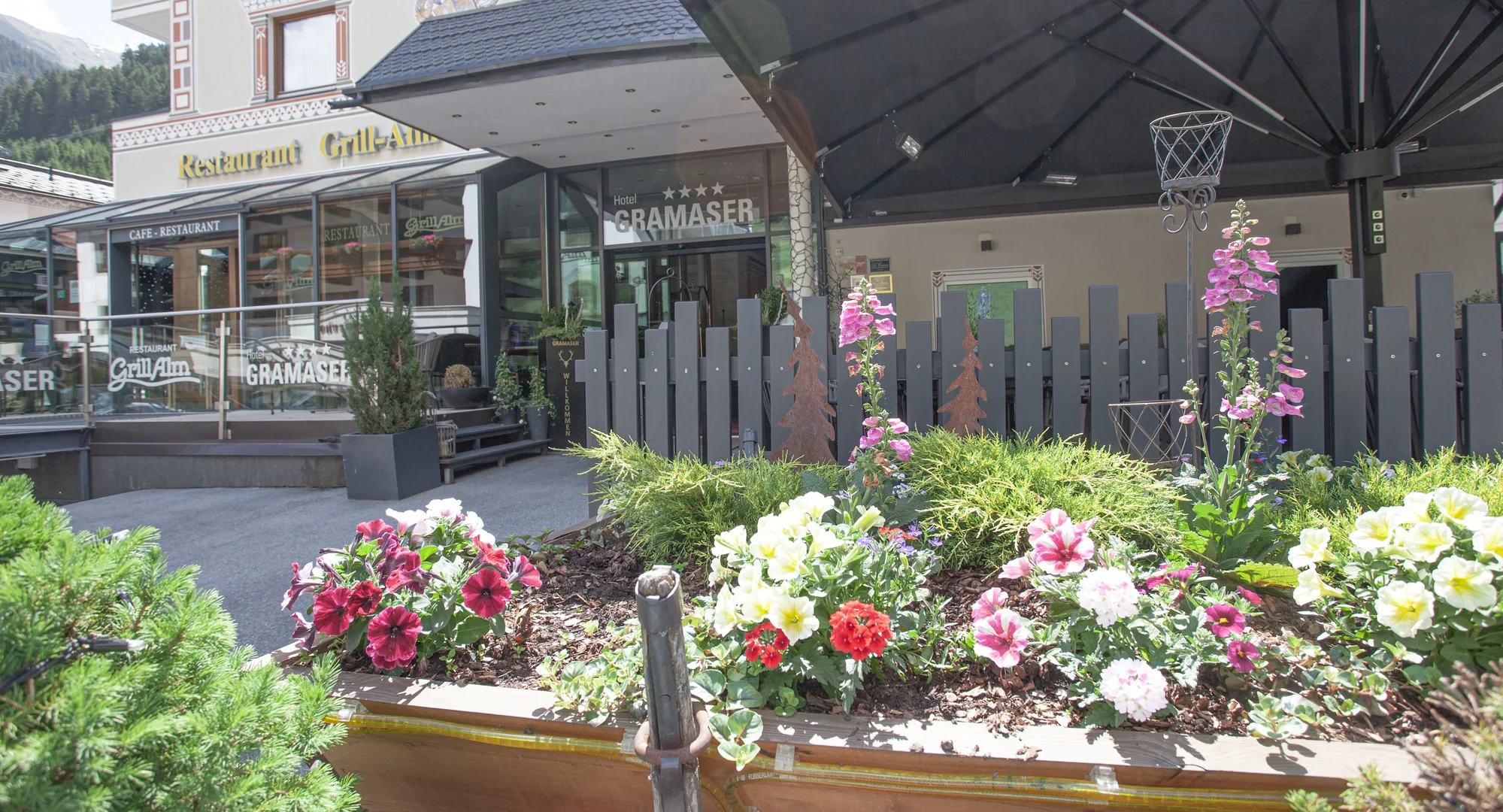 Blühende Blumenkästen vor dem Hotel Gramaser und dem Restaurant Grill-Alm mit Holzzaun und Sonnenschirm.