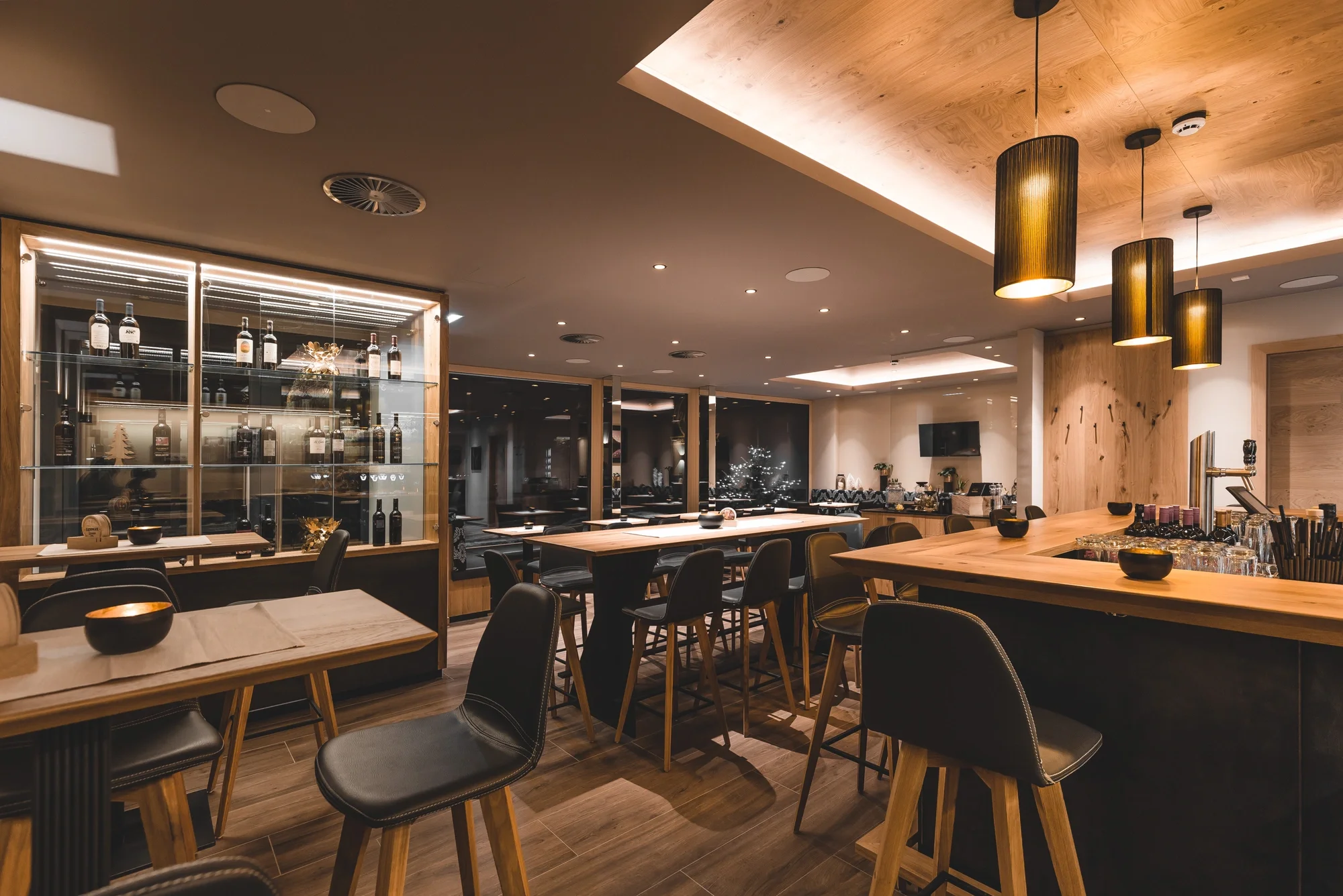 Modernes Restaurant oder Bar mit Holzmöbeln, Weinflaschen in Vitrine, langen Tischen und Barhockern.