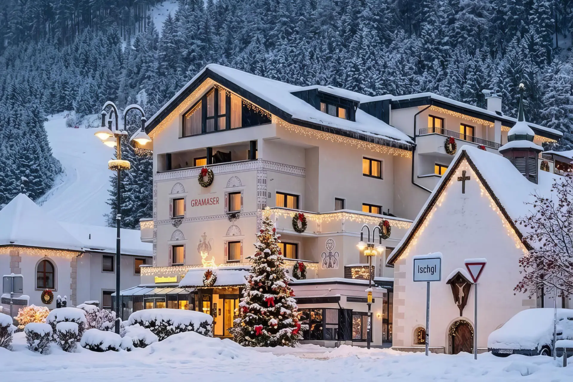 Verschneites Hotel Gramaser in Ischgl mit weihnachtlicher Beleuchtung, Christbaum und kleiner Kapelle im Winter.