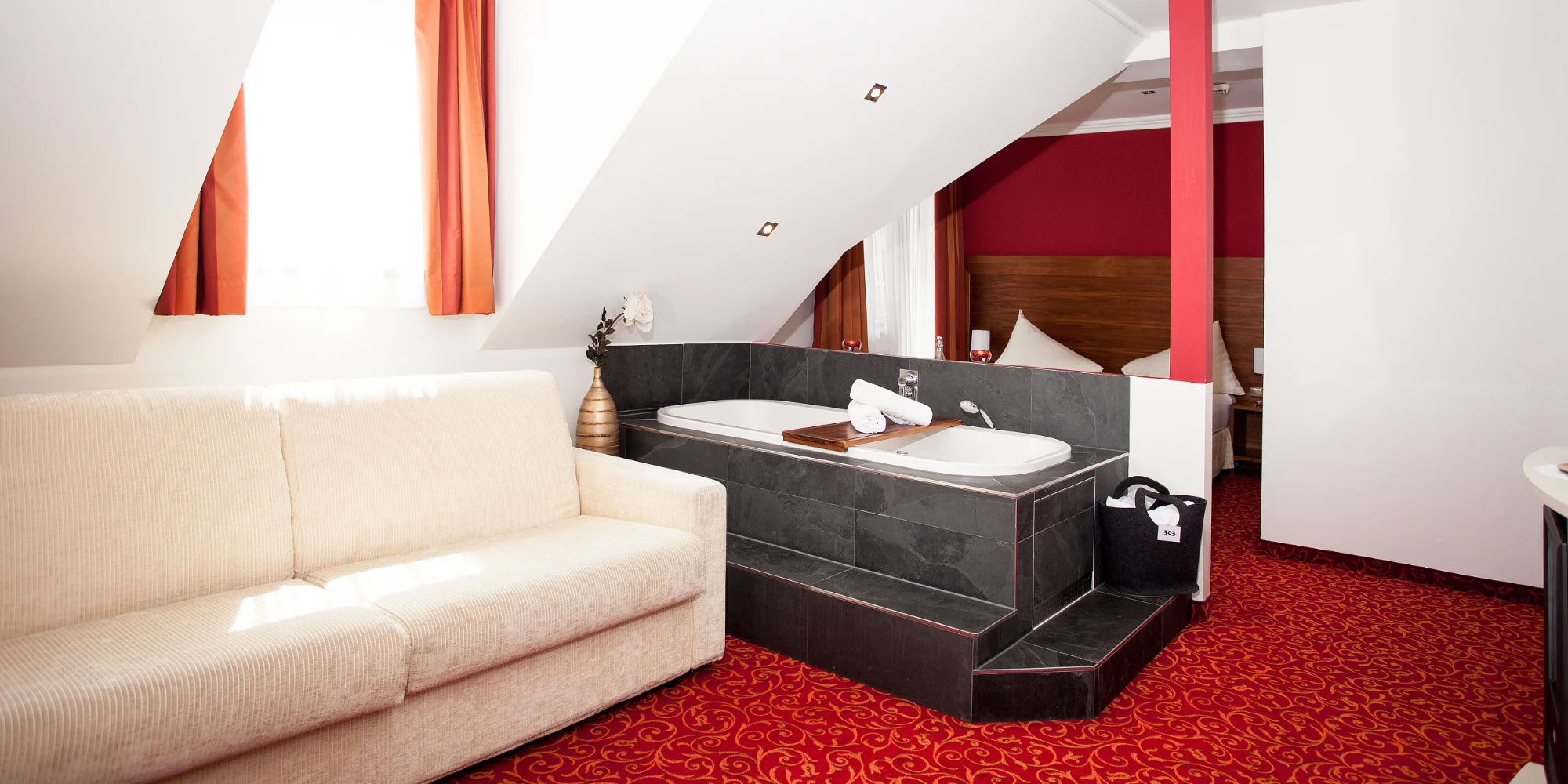 A luxurious hotel room with a red carpet, a bath on a platform, a beige sofa and a view of the bed.